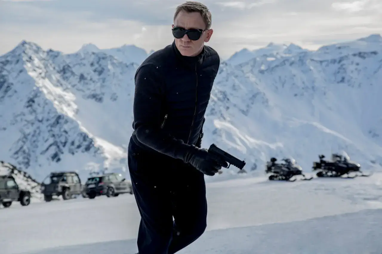 Daniel Craig w okularach przeciwsłonecznych, z pistoletem w dłoni, w śnieżnych górach. To scena, gdzie nie czas umierać.