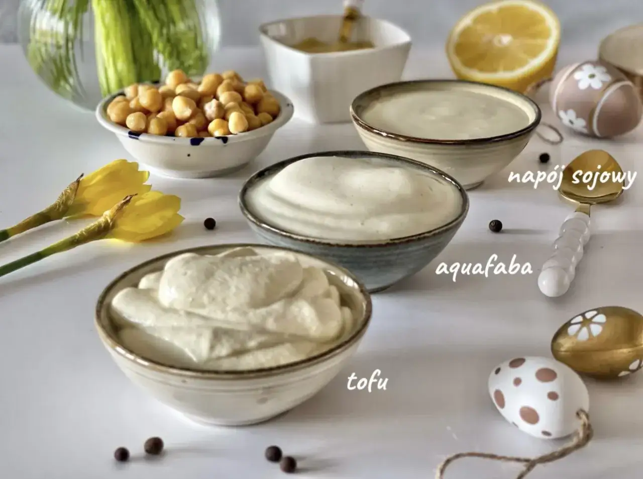 Trzy miseczki z kremowymi, białymi sosami: tofu, aquafaba i napój sojowy. Obok miseczka ciecierzycy, cytryna i ozdobne jajka. Idealne składniki na wegański majonez.