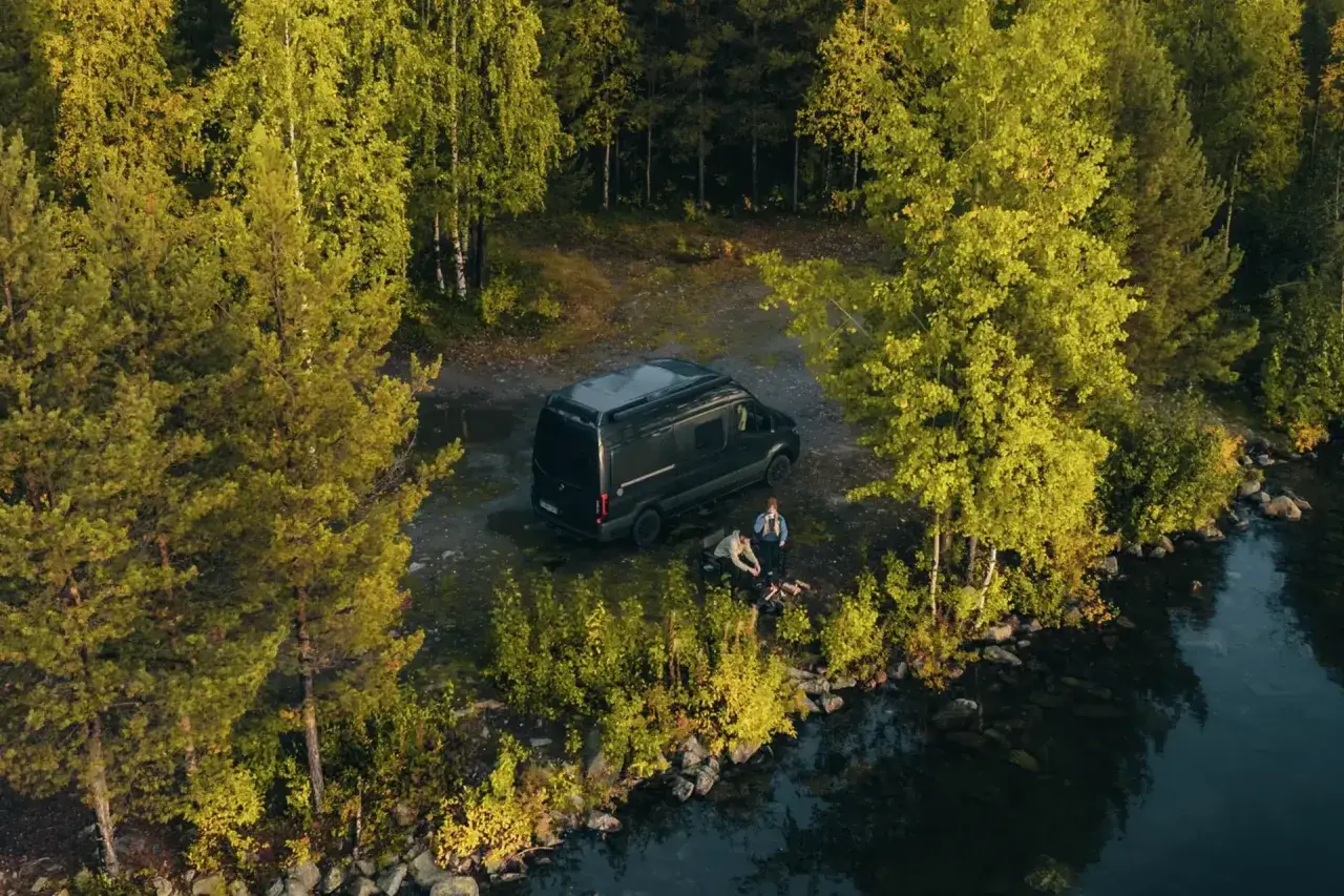 Två personer njuter av van life vid en sjö, omgivna av höstfärgad skog.