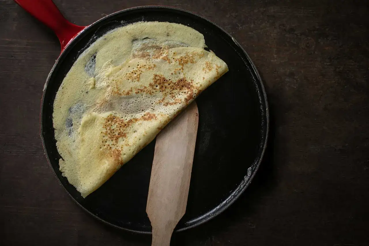 Ein goldbrauner Crêpe wird in einer Pfanne mit einem Holzspatel gefaltet. Perfekt für ein einfaches Crêpe Rezept.