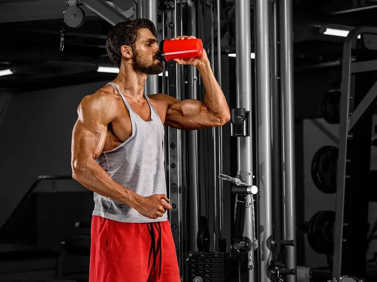 Hombre musculoso bebe de un bidón rojo en el gimnasio, un ejemplo de como tomar proteína para recuperarse.
