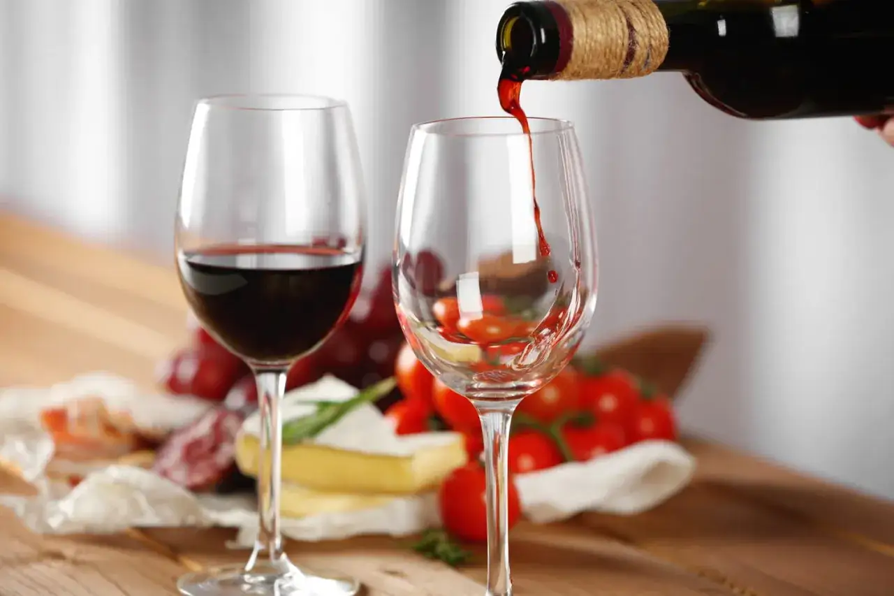 Un verre de vin rouge est servi, à côté d'un autre déjà rempli. Des grappes de raisin, des tomates et du fromage complètent cette scène gourmande de vin au verre.