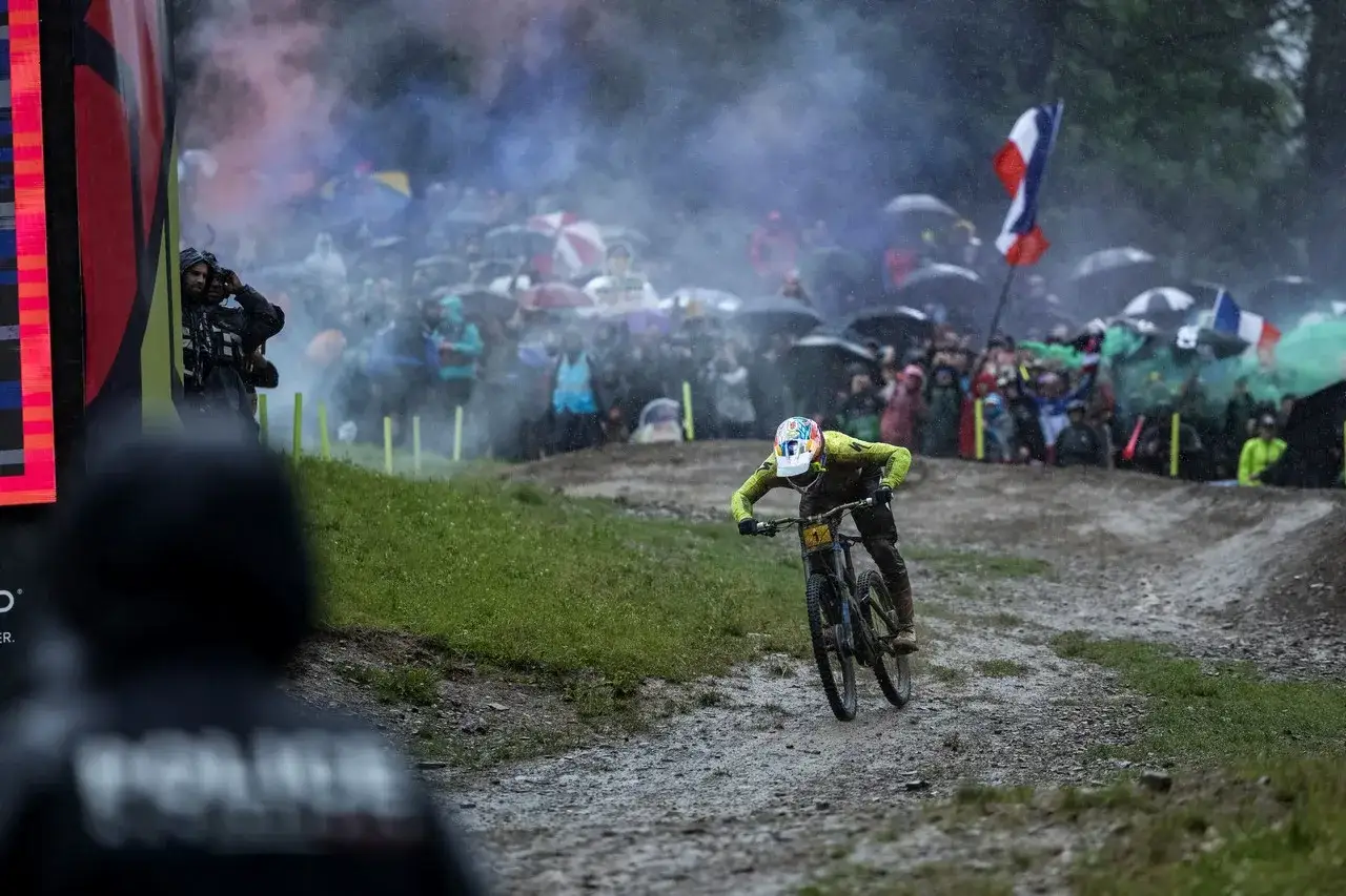 Kolarz MTB w kasku zjeżdża po błotnistym torze, podczas gdy tłum kibiców z parasolami i flagami obserwuje zawody.