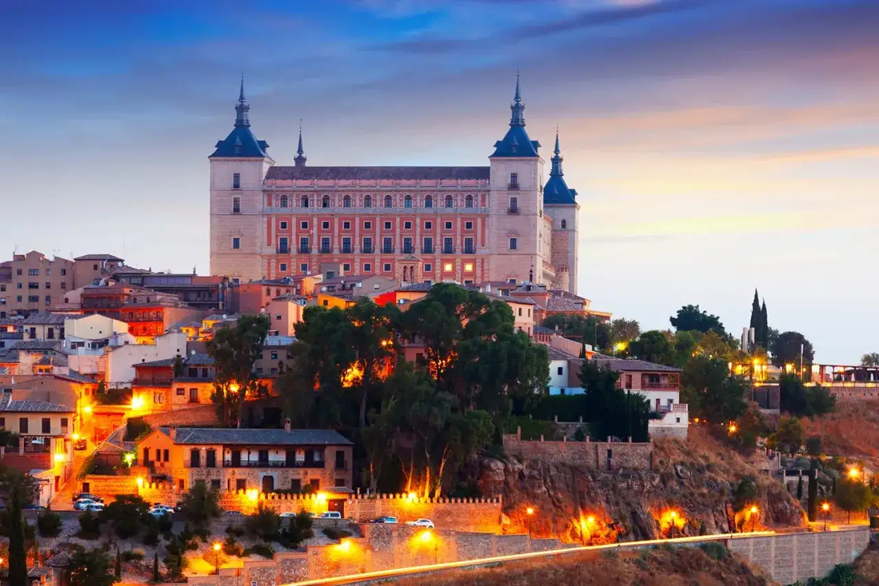 Widok na zabytkowe miasto Toledo, Hiszpania, o zmierzchu. Imponujący Alcázar góruje nad wzgórzem, a uliczki rozświetlają ciepłe światła.
