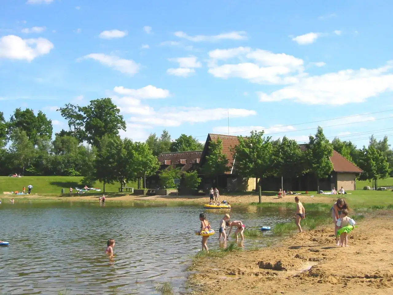 Sonniger Tag am badesee fredenbeck. Kinder planschen im Wasser und am Sandstrand, im Hintergrund Geb&auml;ude und B&auml;ume.