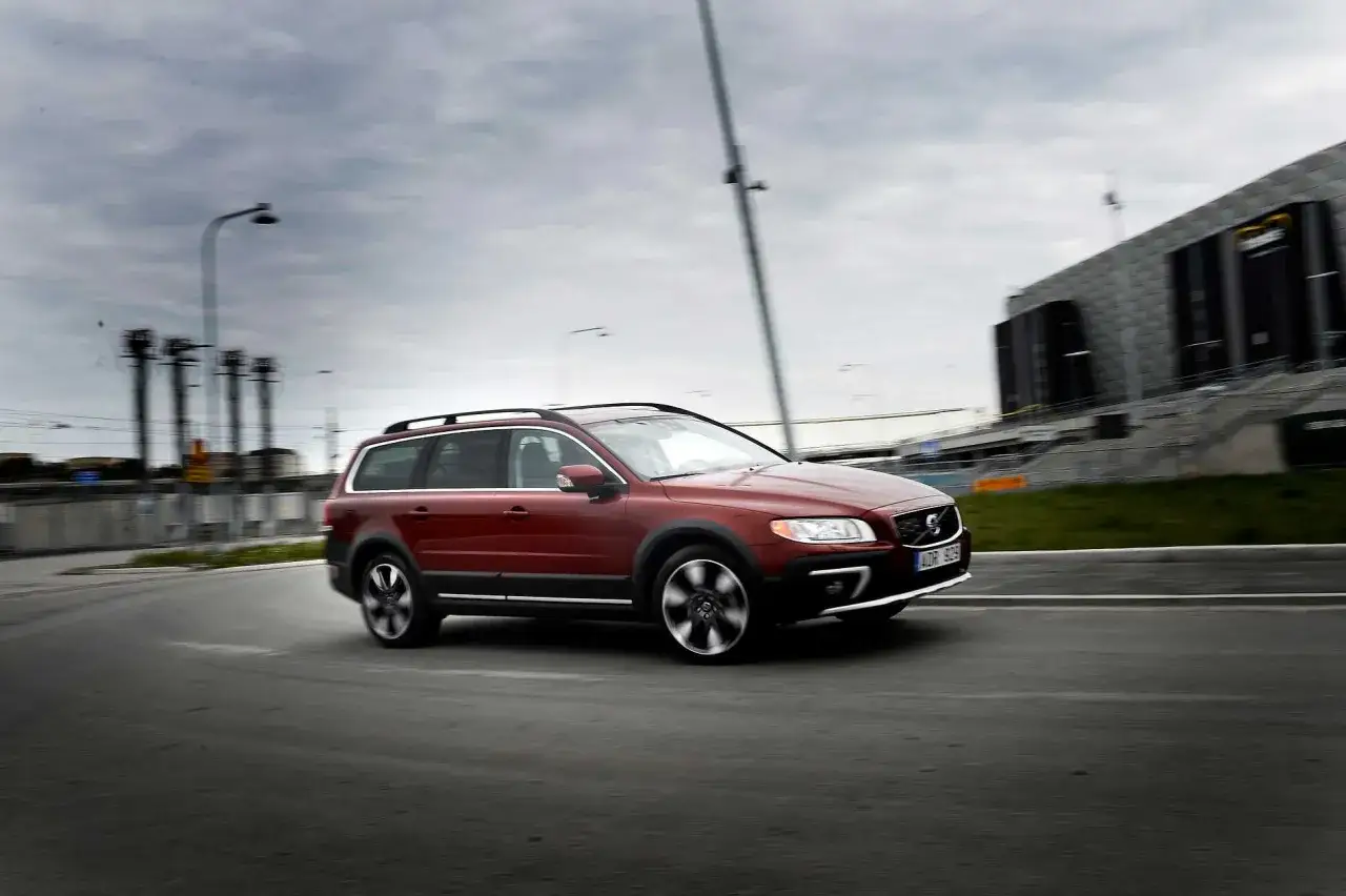 R&ouml;d Volvo XC70 2016 tar en kurva, kanske med ett problem. I bakgrunden syns en modern byggnad och en gr&aring; himmel.