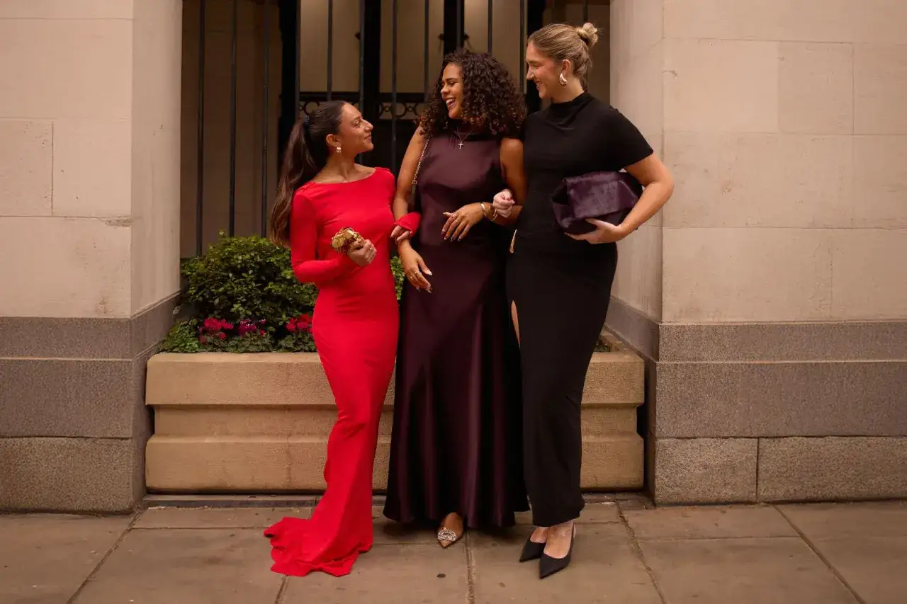 Three women in elegant gowns&mdash;red, plum, and black&mdash;discussing the best colors to wear to a wedding.