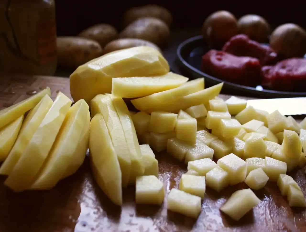 Skalade potatisar, skurna i klyftor och t&auml;rningar, redo f&ouml;r att bli h&auml;rligt r&aring;stekt potatis. I bakgrunden syns fler potatisar och k&ouml;tt.