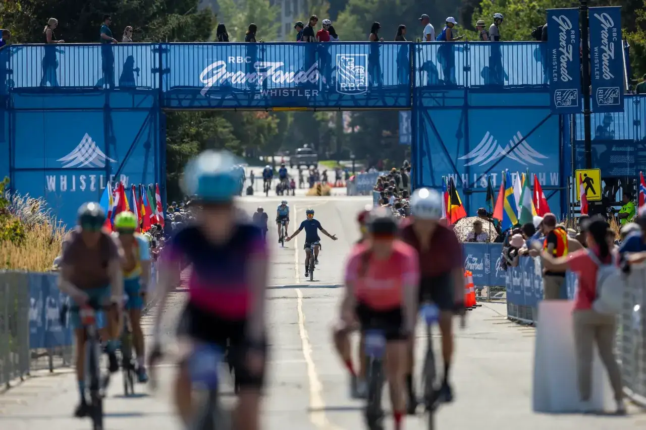 Cyclists race under a blue archway at the RBC Gran Fondo Whistler. Spectators cheer, showcasing what a sportive event is all about.