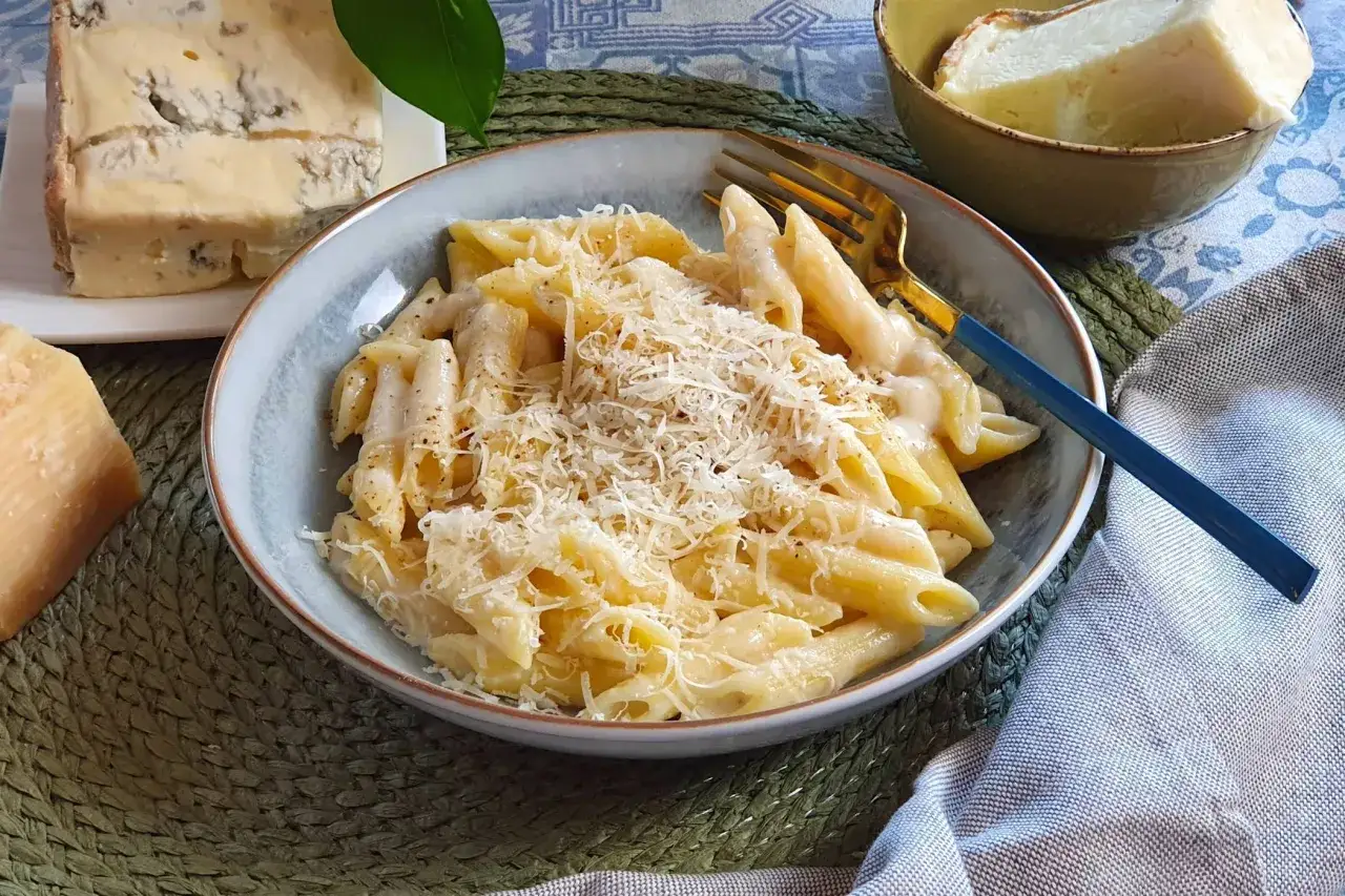 Piatto di penne cremose, condite con una ricca salsa ai 4 formaggi, guarnite con parmigiano grattugiato. Accanto, pezzi di formaggio fresco e stagionato.
