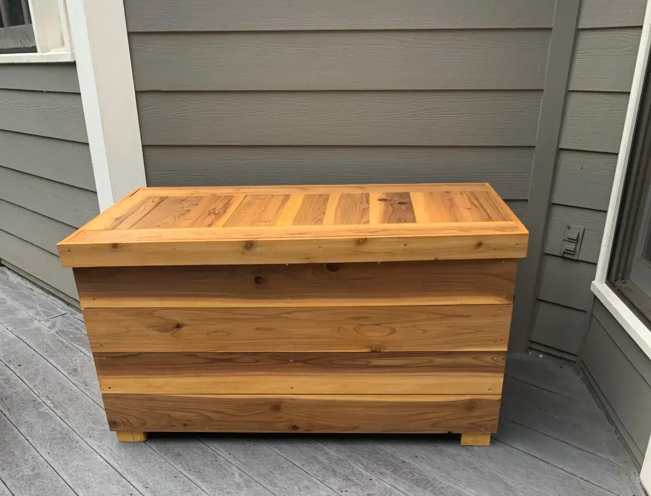 A beautiful, handcrafted diy deck box made of natural wood sits on a grey deck. Its slatted design and warm tones complement the surrounding house siding.