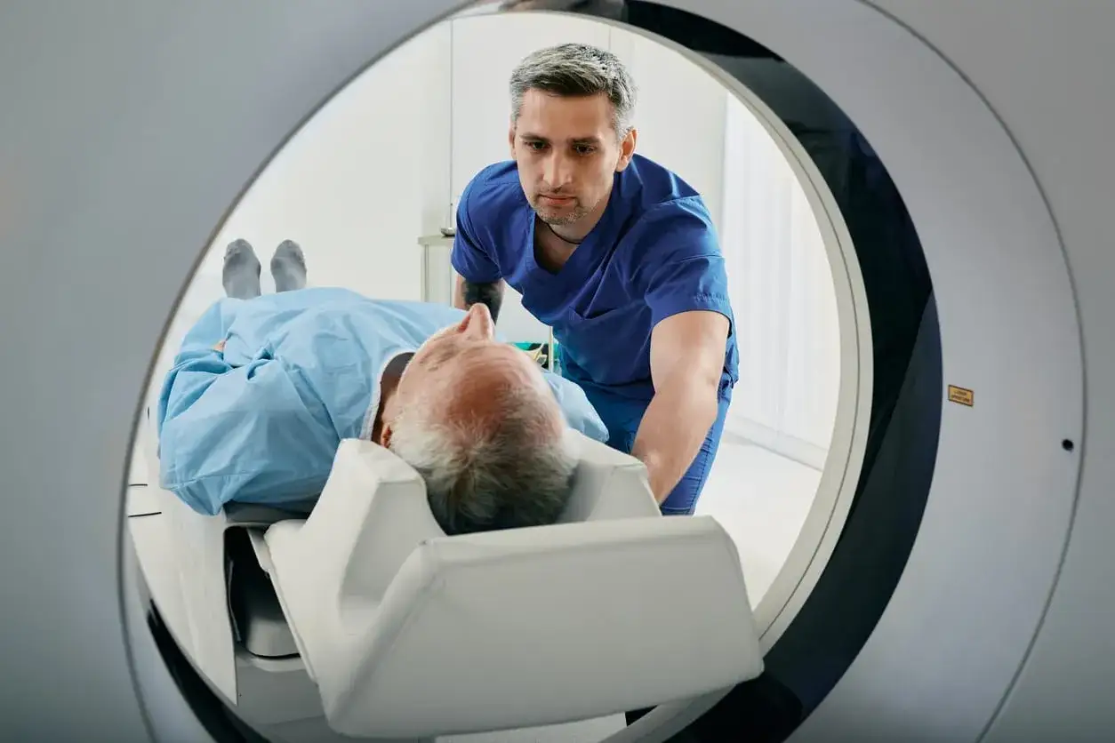 Un patient &acirc;g&eacute; est plac&eacute; dans un scanner TDM. Le technicien, en blouse bleue, le positionne pour l'examen. TDM c'est quoi ? C'est une technologie d'imagerie m&eacute;dicale.
