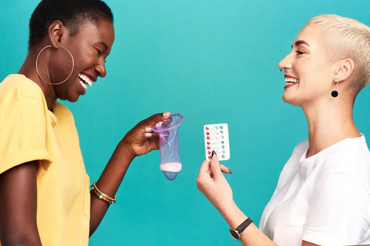 Dos mujeres r&iacute;en mientras una muestra un cond&oacute;n y la otra pastillas anticonceptivas, discutiendo c&oacute;mo adelantar la regla.