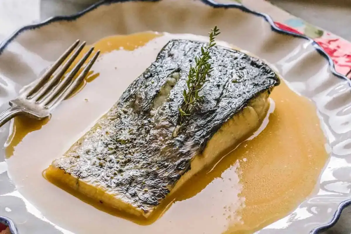 Filete de pescado crujiente bañado en una cremosa salsa beurre blanc, adornado con una ramita de romero.