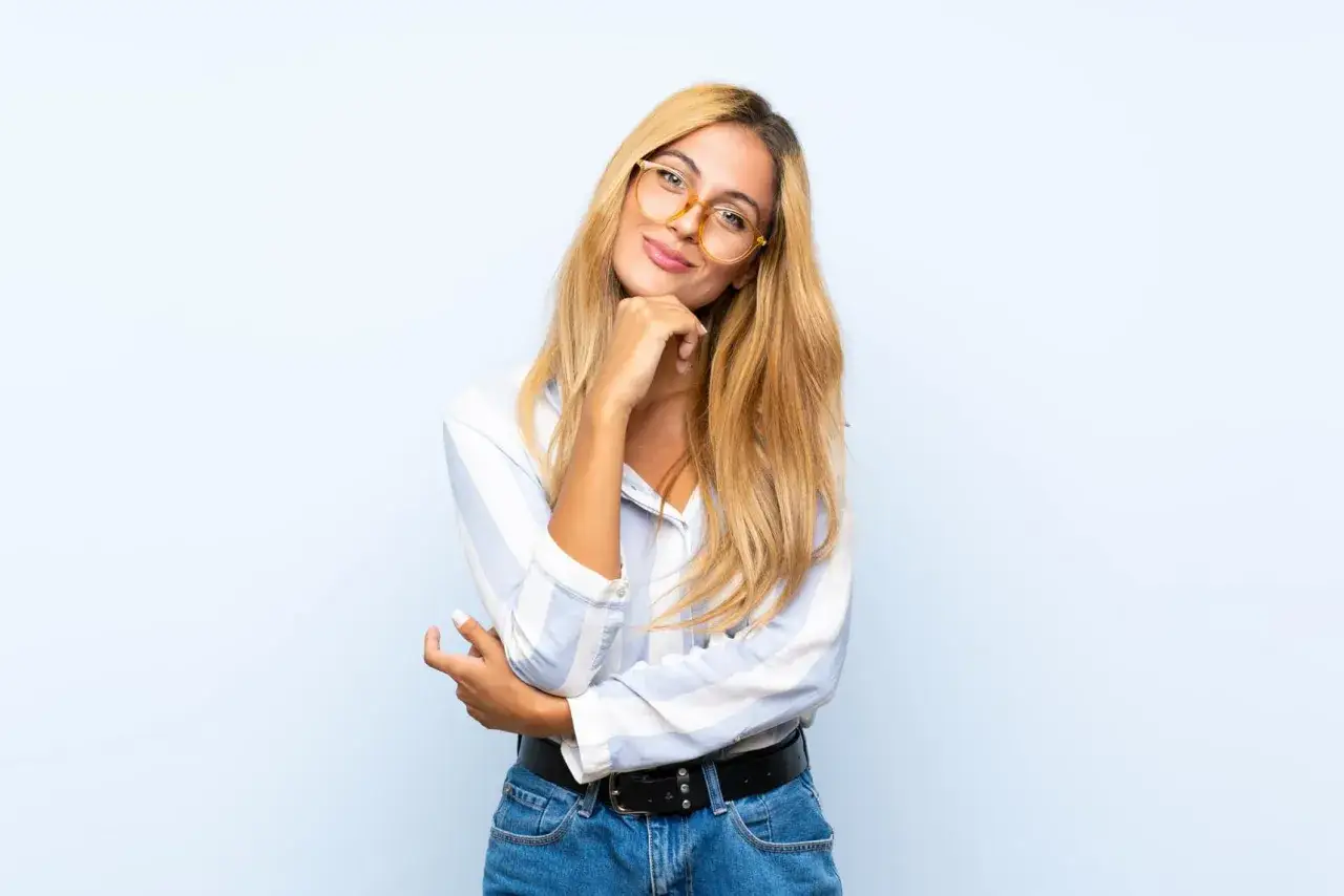 Uśmiechnięta blondynka w okularach dla blondynek, w koszuli w paski i dżinsach, z ręką na podbródku.