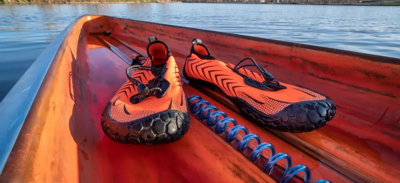 Pomarańczowe buty wodne z czarnymi akcentami, idealne jako buty na kajaki, leżą w kajaku na tle spokojnej wody.