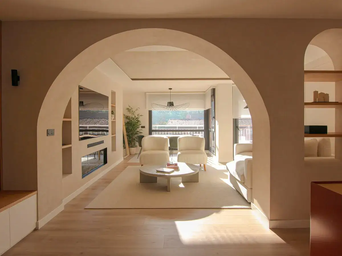 Amplio y luminoso sal&oacute;n con entrada recibidor integrado. Dos sillones blancos, mesa de centro redonda y sof&aacute;, bajo una l&aacute;mpara de dise&ntilde;o.