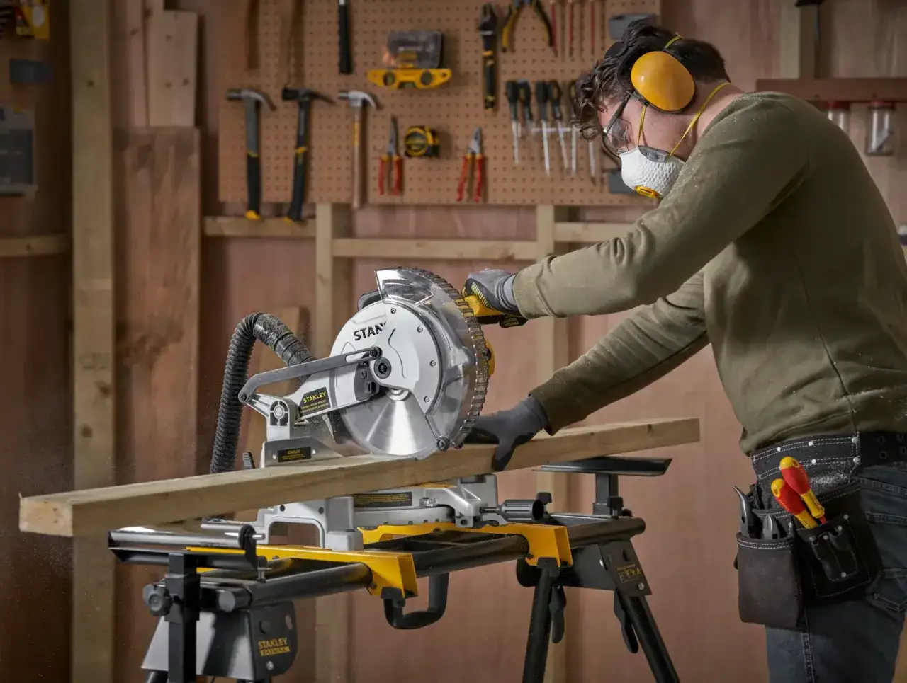 Un bricoleur utilise une scie à onglet pour couper du bois, protégé par des lunettes, un masque et des cache-oreilles. Il montre comment utiliser une scie à onglet en toute sécurité.