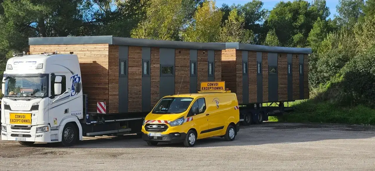 Un camion blanc tracte une longue structure en bois et métal. Une camionnette jaune, signalant un convoi exceptionnel, suit de près.