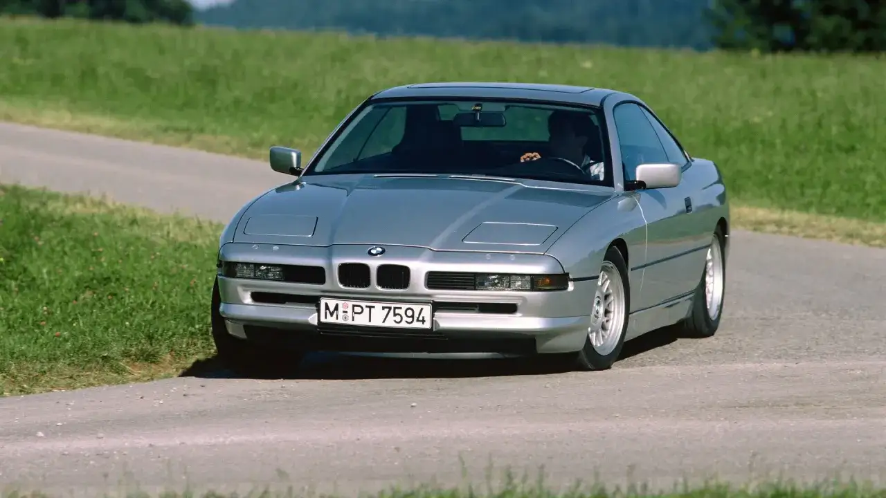 Un elegante BMW E31 plateado recorre un camino rural rodeado de exuberante vegetaci&oacute;n.