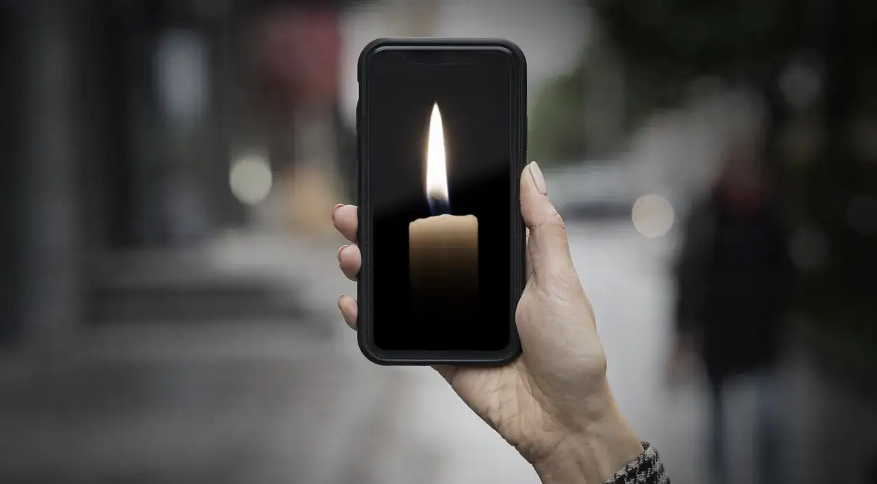 Dłoń trzyma telefon z płonącą świecą na ekranie, symbolizującą pamięć o zmarłej osobie.