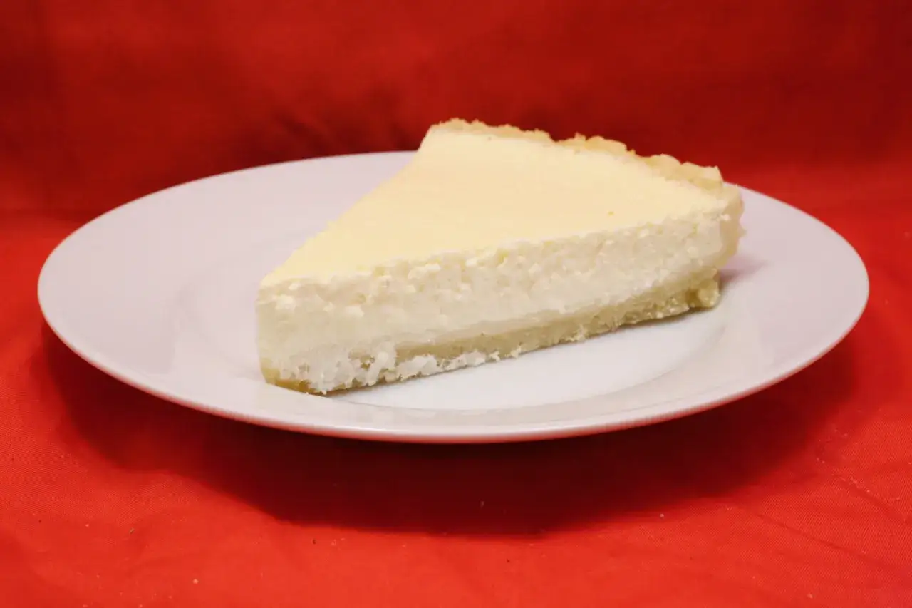 Ein St&uuml;ck cremiger K&auml;sekuchen mit einem Hauch von saurer Sahne auf einem wei&szlig;en Teller vor rotem Hintergrund.