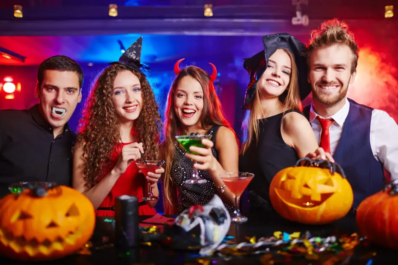 Glada vuxna i halloweenkostymer med pumpor och drinkar. En vampyr, en h&auml;xa, en dj&auml;vul och en man i kostym njuter av festen.