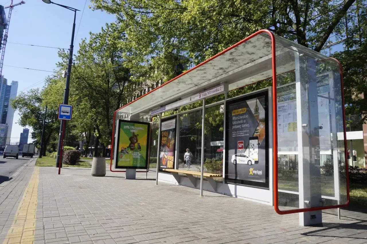 Nowoczesne, przeszklone przystanki autobusowe z czerwonymi ramami. Na jednym z nich reklama mytaxi.
