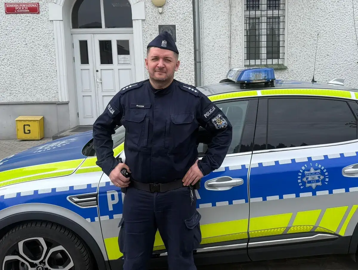 Policjant w mundurze stoi obok radiowozu. Jego postawa sugeruje gotowość do działania, a obecność policyjnego pojazdu podkreśla jego rolę w zapewnianiu dozoru policyjnego.
