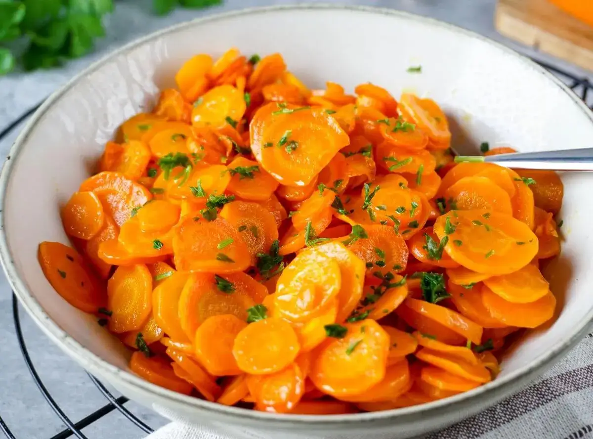 Schüssel mit leuchtend orangefarbenen, in Scheiben geschnittenen Karotten, garniert mit frischer Petersilie. Perfekt gekochte Karotten, die Lust auf mehr machen.