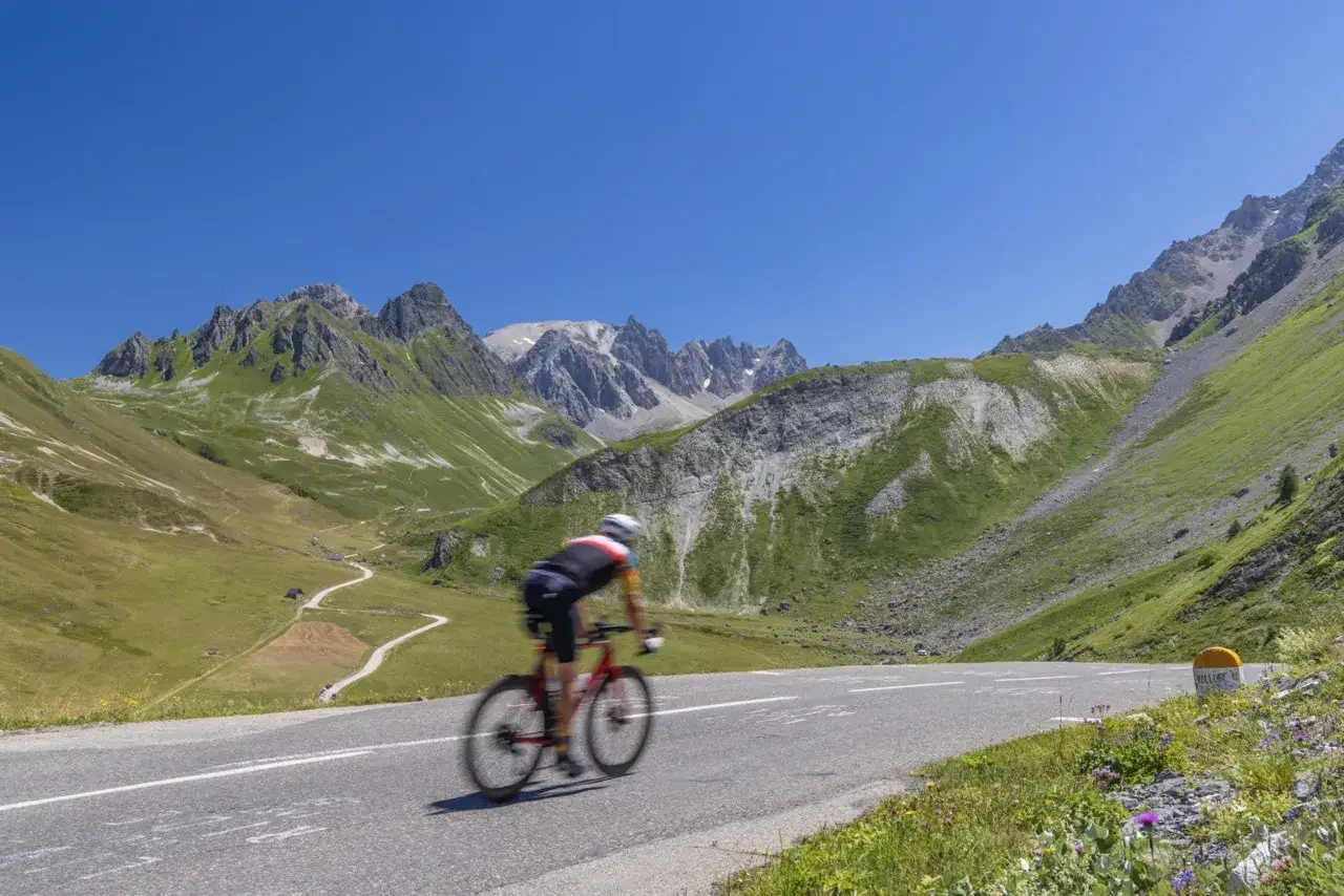 Cyclist Isaac Del Toro powers up a winding mountain road, surrounded by lush green slopes and jagged peaks under a clear blue sky.