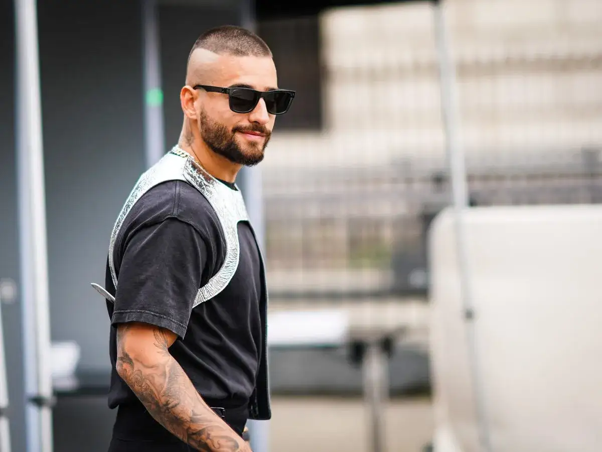 Hombre con un moderno buzz cut largo, gafas de sol y un llamativo arn&eacute;s plateado sobre camiseta negra.
