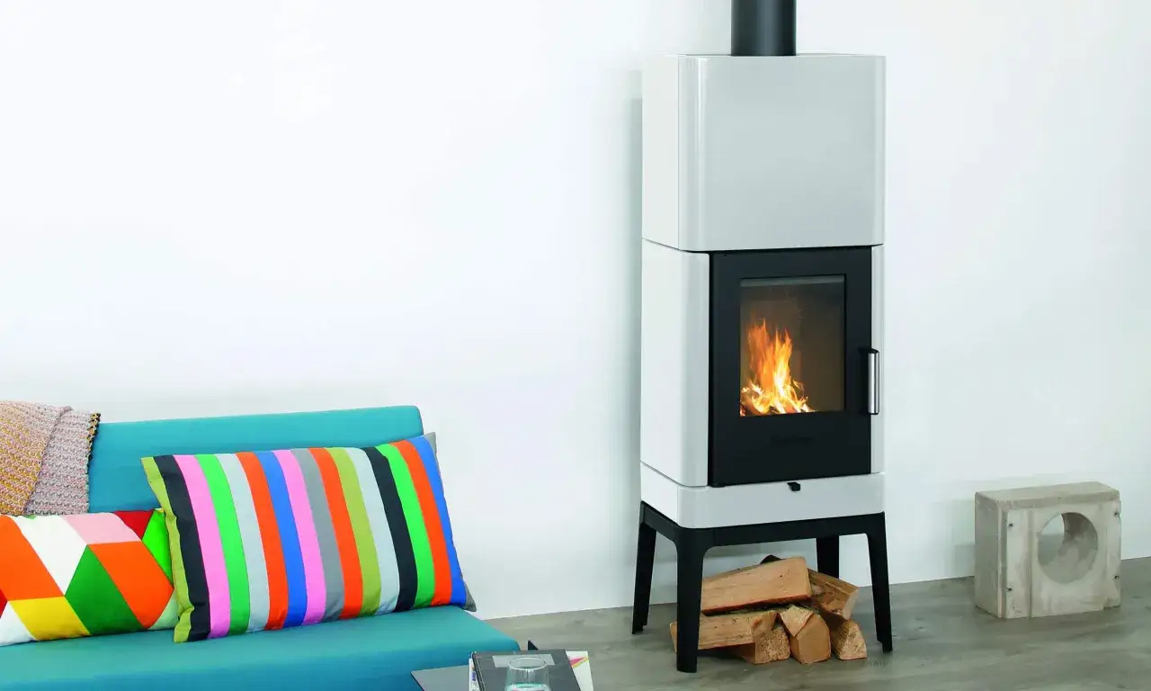 Ein moderner Bullerjan B3 Ofen mit brennendem Feuer neben einem türkisfarbenen Sofa mit bunten Kissen.
