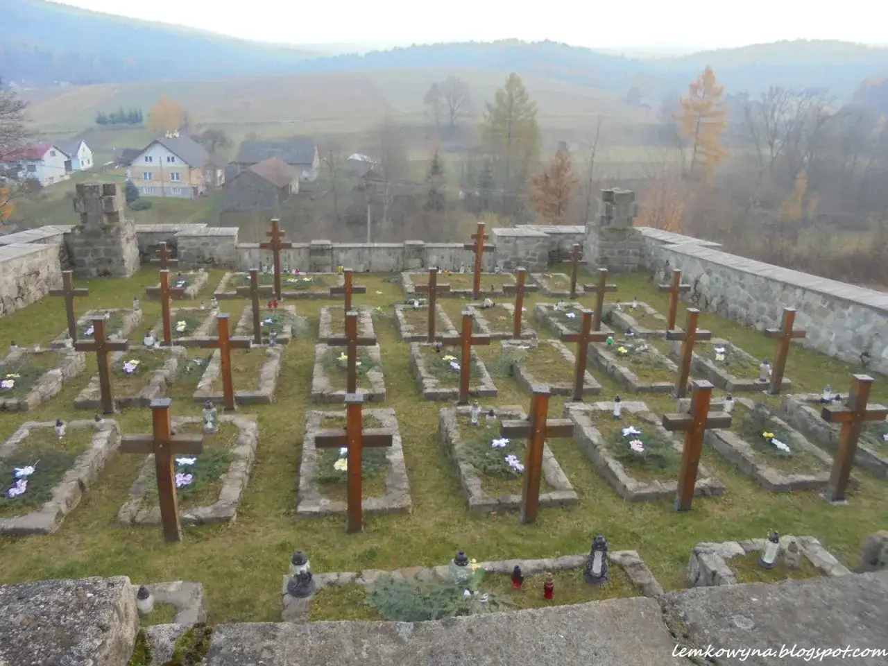 Cmentarz z drewnianymi krzyżami i kamiennymi nagrobkami, otoczony murem. W tle widać wieś i wzgórza. To miejsce wiecznego spoczynku, gdzie każdy krzyż to symbol ożenna.