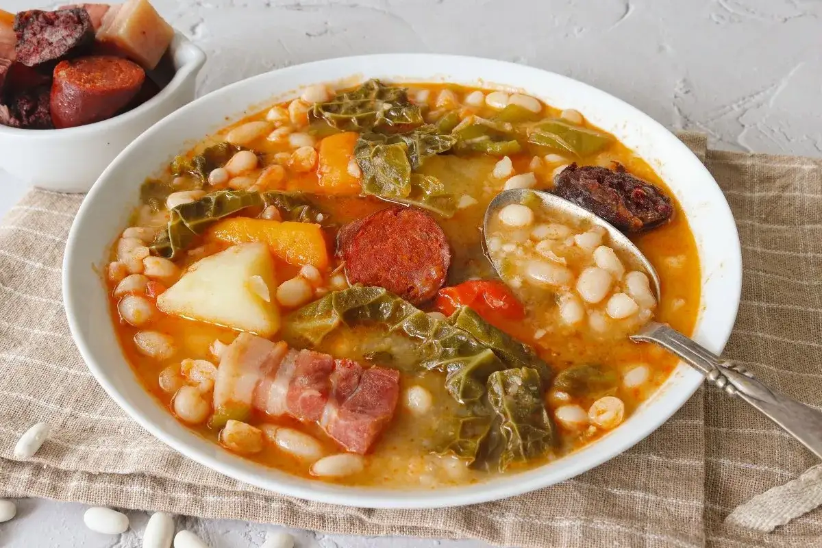 Un reconfortante cocido monta&ntilde;&eacute;s, con alubias blancas, chorizo, morcilla, tocino y verduras, servido en un plato blanco.