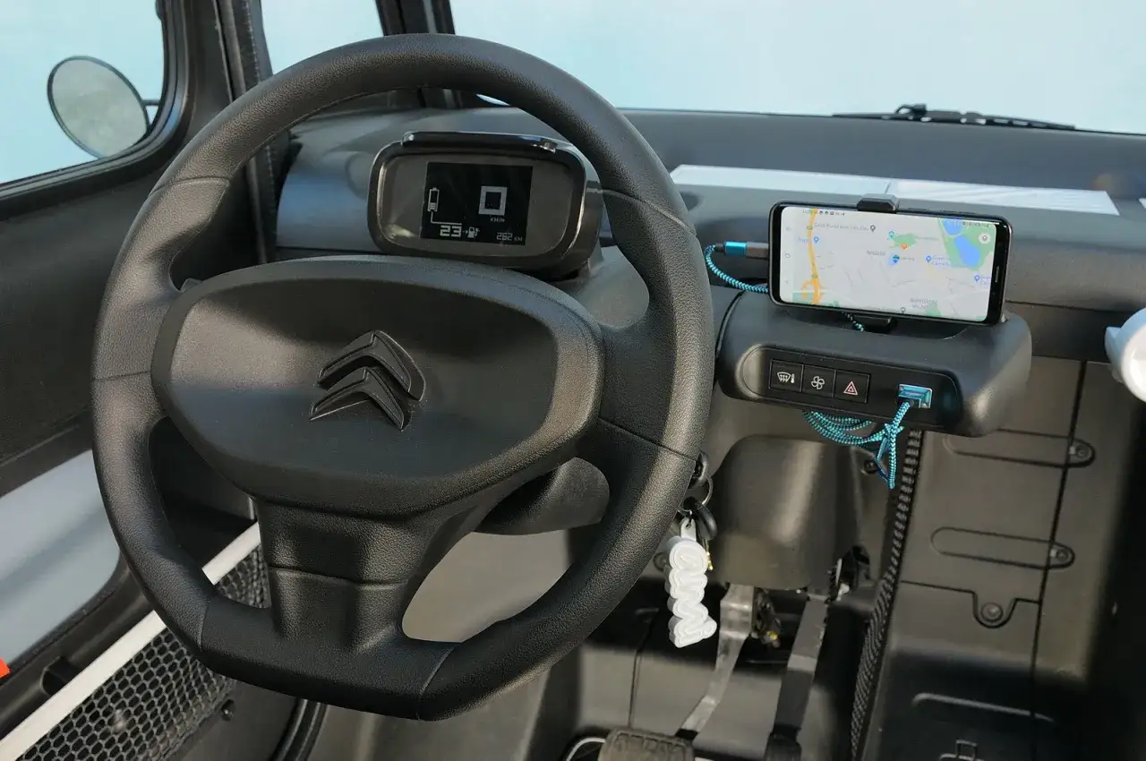 Interno Citroen Ami prova: volante, cruscotto digitale, smartphone con navigatore e cavi di ricarica.