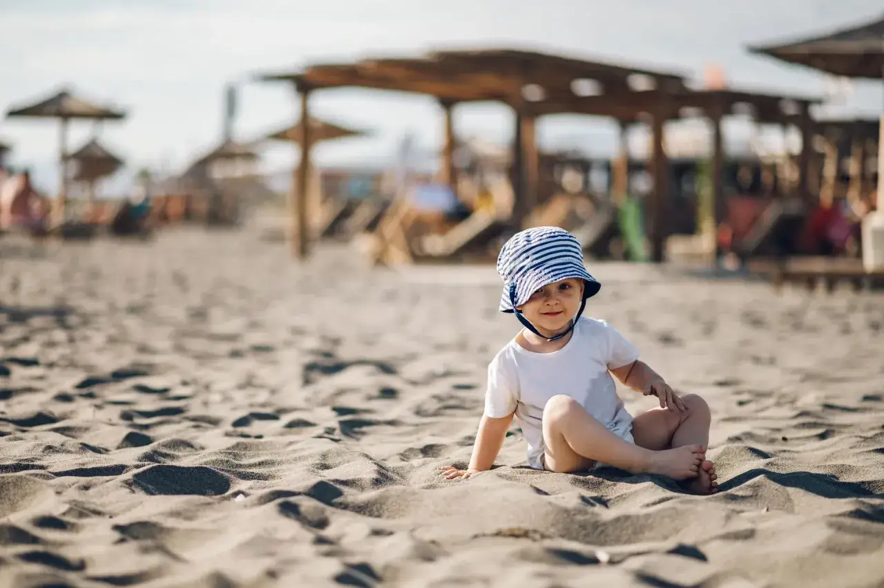 Uroczy maluch w kapelusiku na plaży. Idealne miejsce, gdzie jechać z rocznym dzieckiem na wakacje.