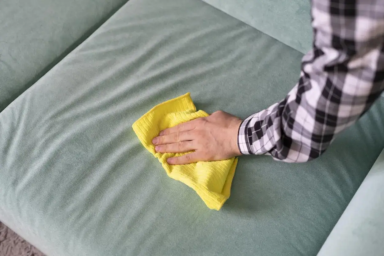 Main d'une personne nettoyant un canap&eacute; en tissu avec un chiffon jaune. Une astuce pour nettoyer canap&eacute; tissu vinaigre blanc bicarbonate.