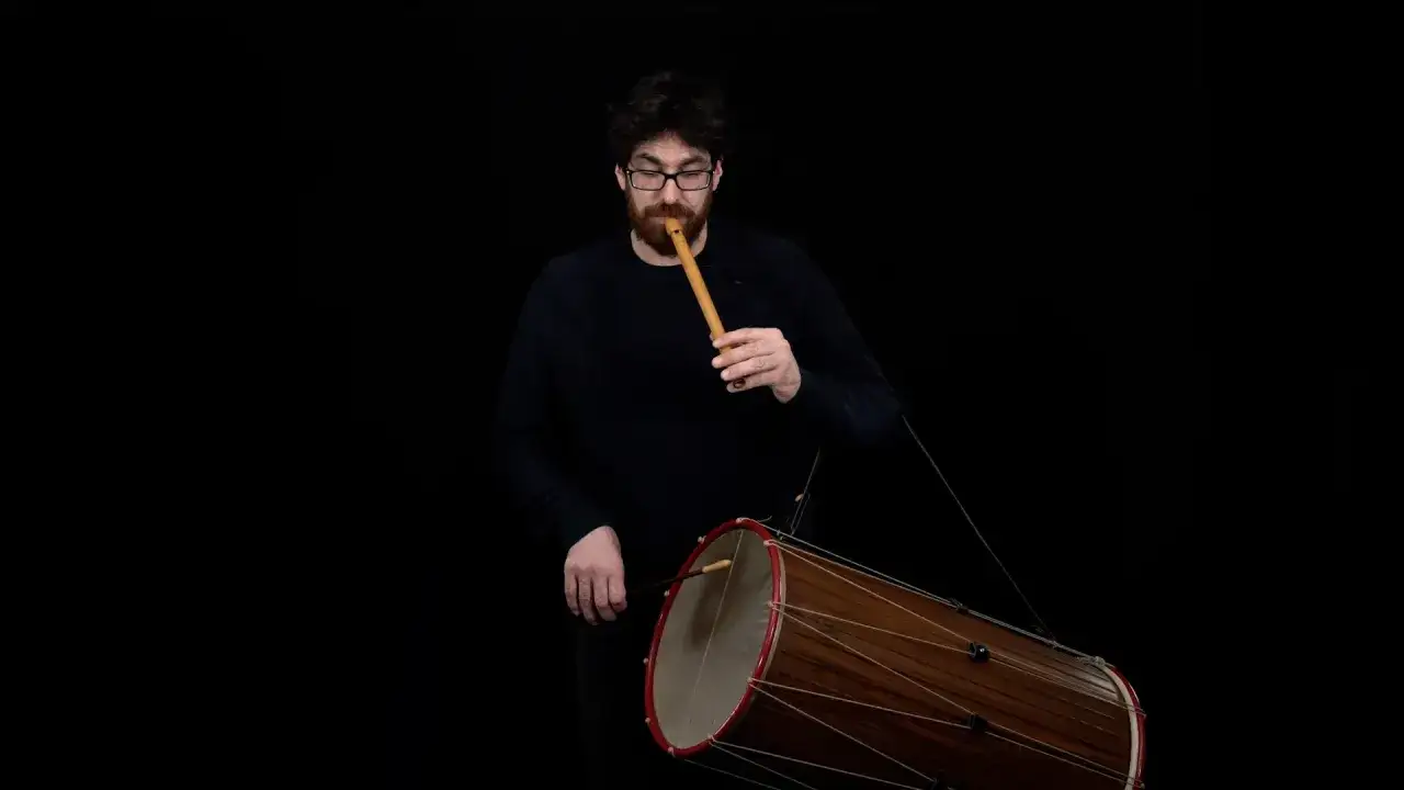 Un homme joue d'un instrument bois, une fl&ucirc;te, tout en frappant un grand tambour.