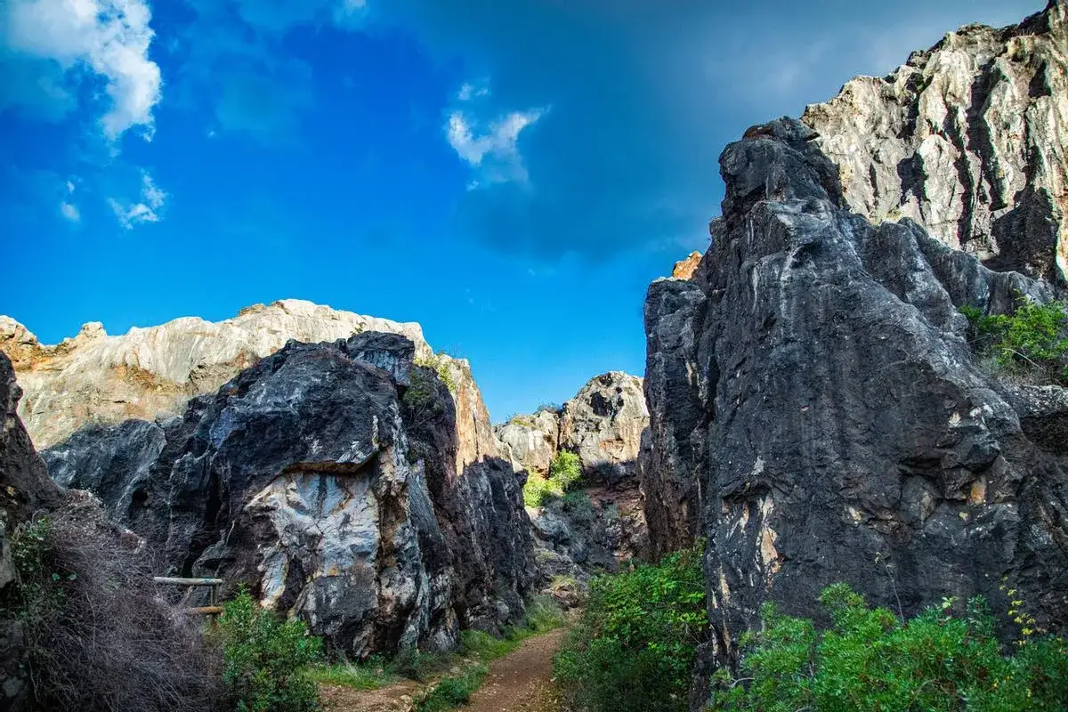 Un sendero serpentea entre imponentes formaciones rocosas de tonos oscuros y claros en el **Cerro del Hierro**, bajo un cielo azul intenso con nubes blancas.