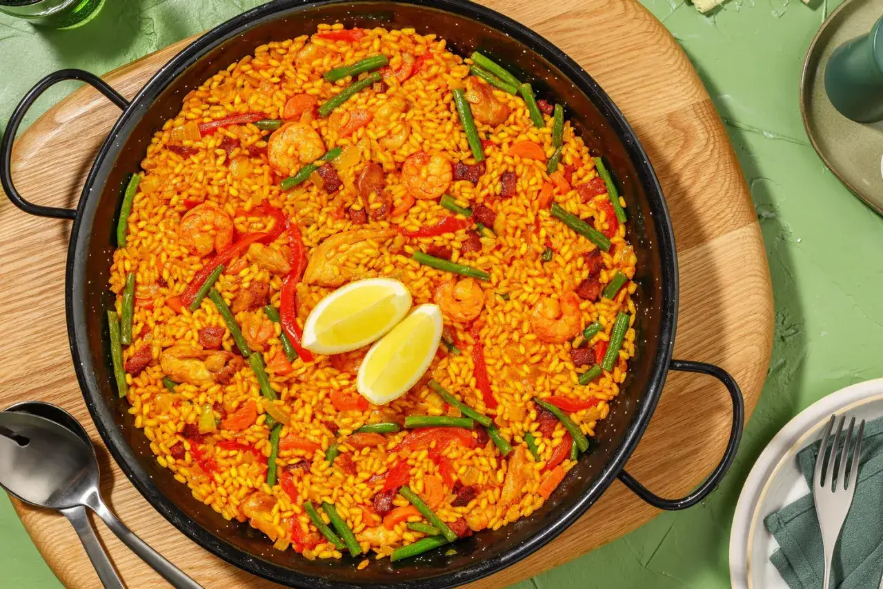 Un plat de riz façon paella, garni de crevettes, poulet, chorizo, poivrons et haricots verts, avec deux quartiers de citron.