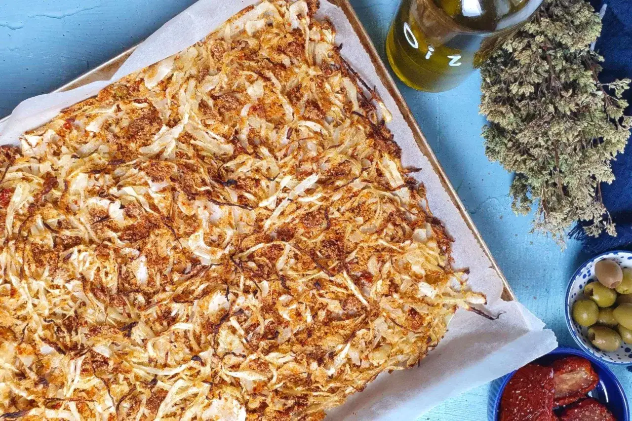 Cavolfiore al forno croccante e dorato, pronto per essere gustato. Accanto, olive verdi, pomodori secchi e un rametto di erbe aromatiche.