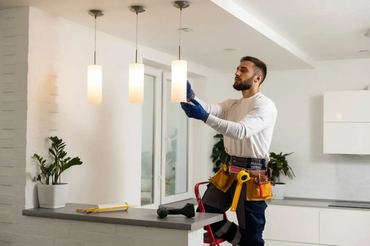 Un **&eacute;lectricien artisan** installe des luminaires modernes. Il est sur un escabeau, portant des gants bleus et une ceinture &agrave; outils.