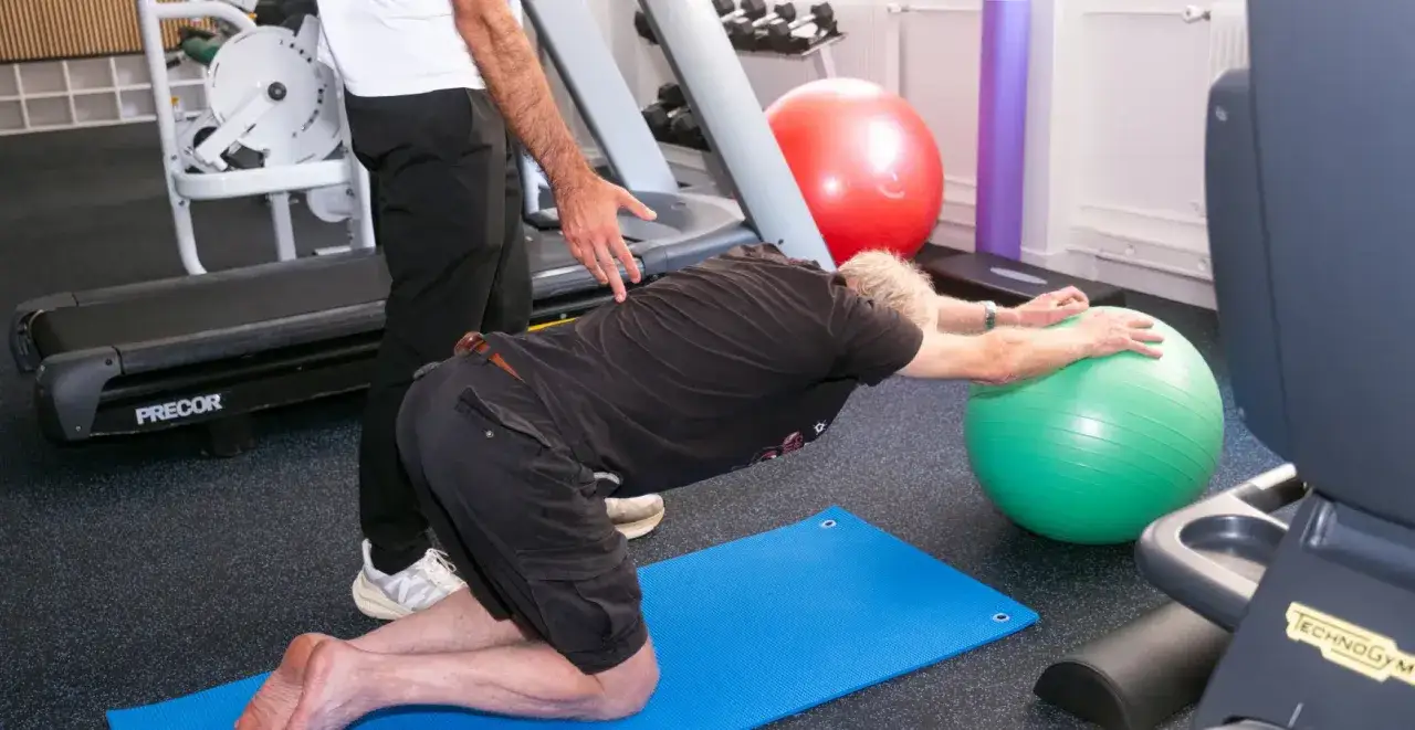 Un homme âgé, souffrant d'arthrose lombaire, s'étire avec l'aide d'un kinésithérapeute sur un tapis bleu, s'appuyant sur un ballon de gym vert.