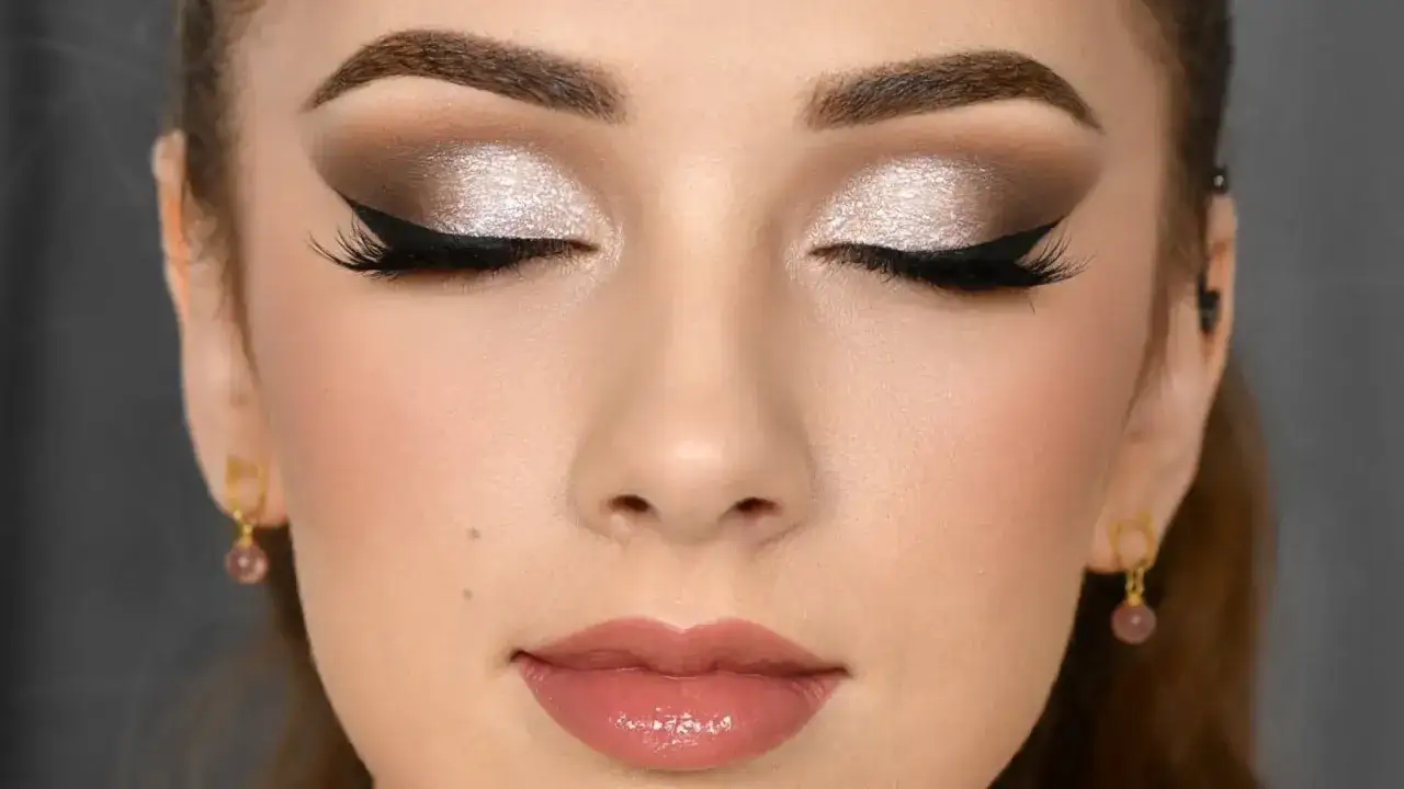 Ojos cerrados con un maquillaje **soft glam** que resalta con sombras plateadas y delineado cat eye. Labios jugosos y pómulos rosados.