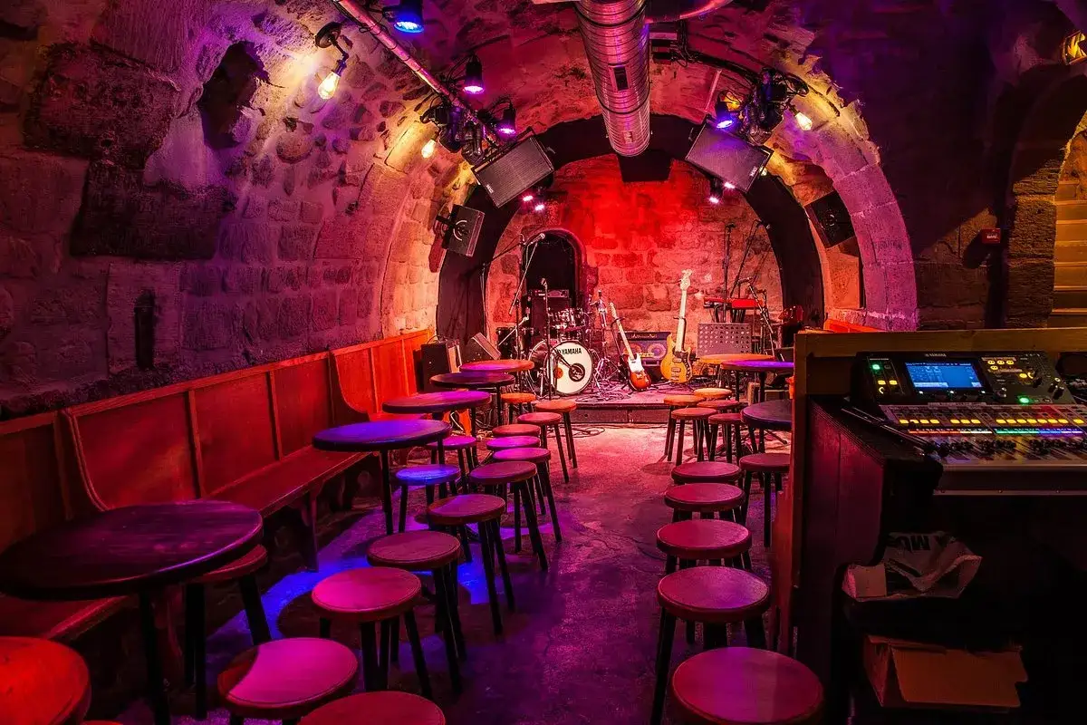 Scène de concert dans le caveau des oubliettes, éclairée par des lumières violettes et rouges. Des tables et des tabourets sont disposés devant la scène où se trouvent des instruments de musique.