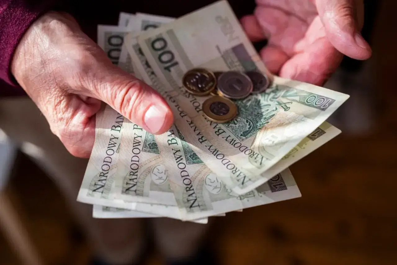 Dłonie trzymają polskie banknoty 100 zł i monety. Może to być wypłata, oszczędności lub środki na staż pracy ZUS.