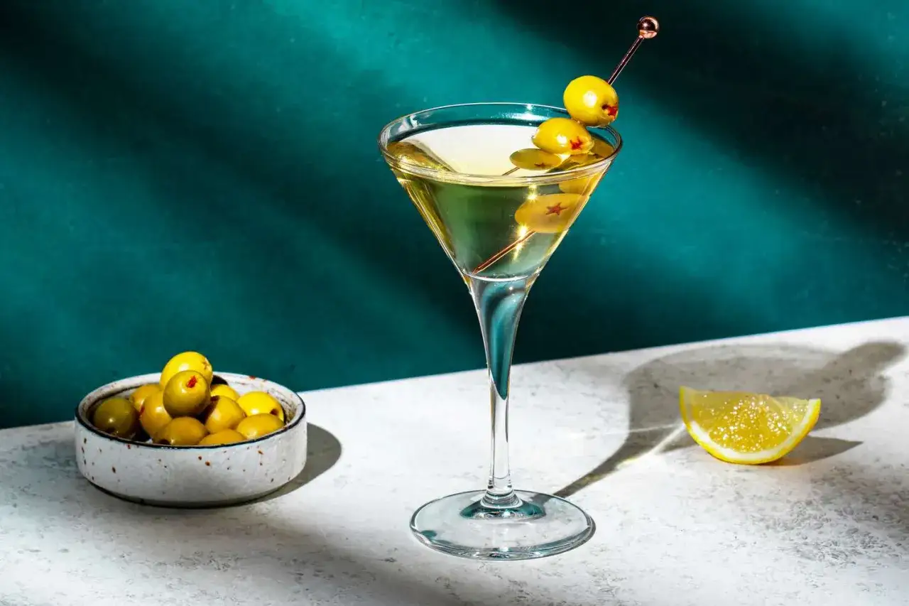 Elegancki martini z wódką, ozdobiony oliwkami na wykałaczce, obok miseczki z oliwkami i plasterka cytryny.