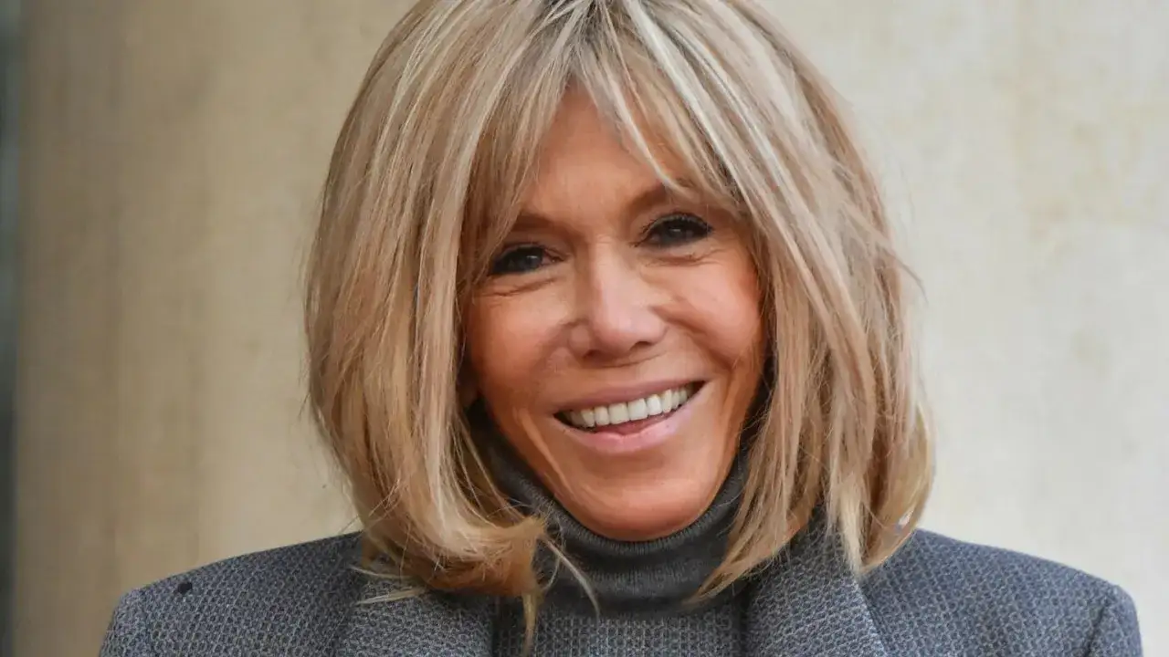 Uśmiechnięta Brigitte Macron z charakterystycznymi blond włosami, w szarym golfie i marynarce.