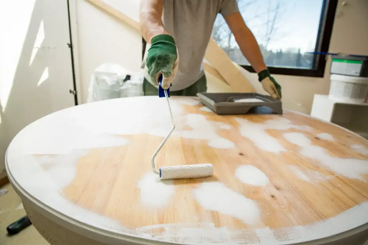 A person in gloves is using a roller to apply white primer to a wooden table, beginning the process of how to refinish a table.